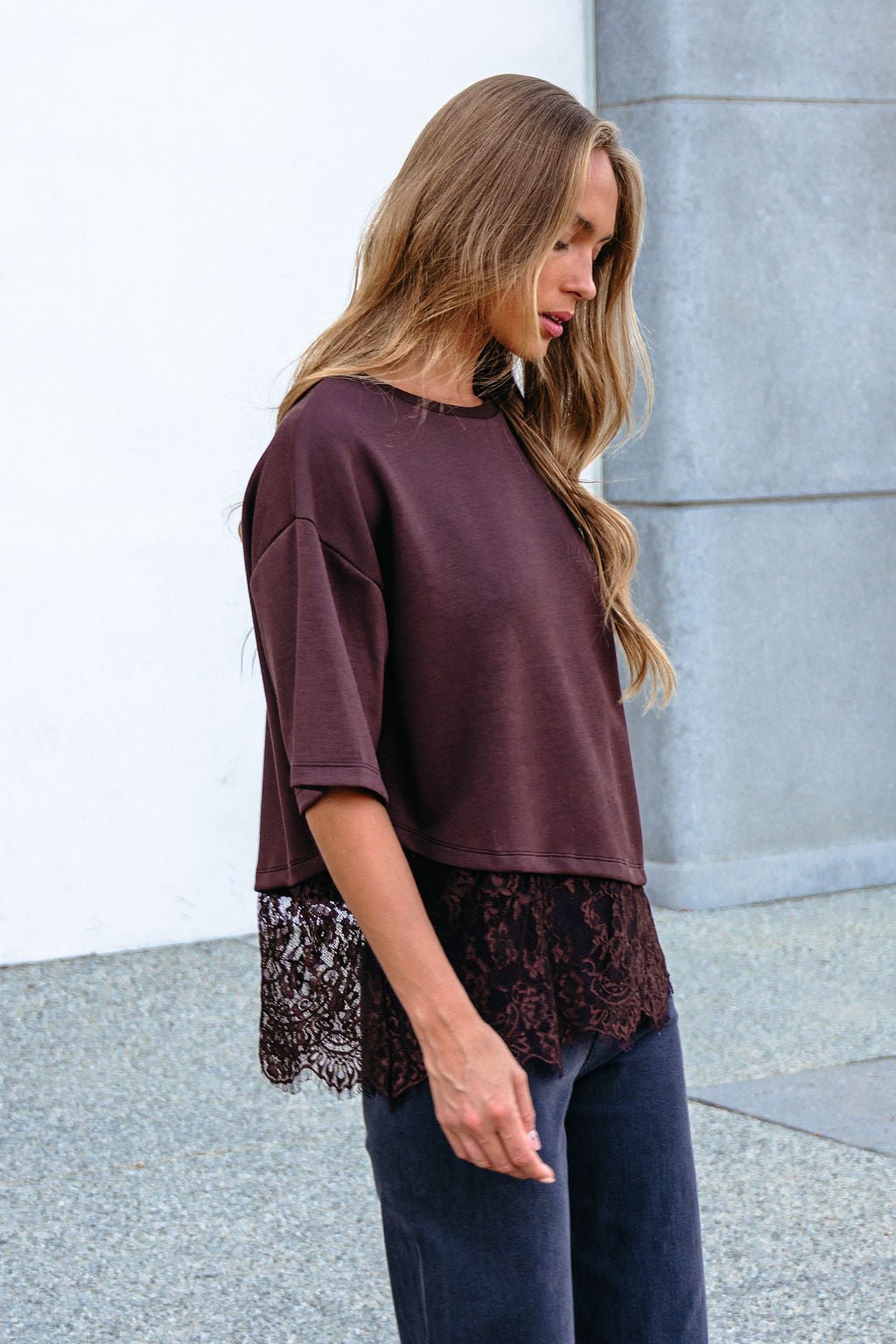 A woman wears a Brown Lace Trim Short Sleeve Top with an oversized fit, standing outside on a sidewalk and looking down.