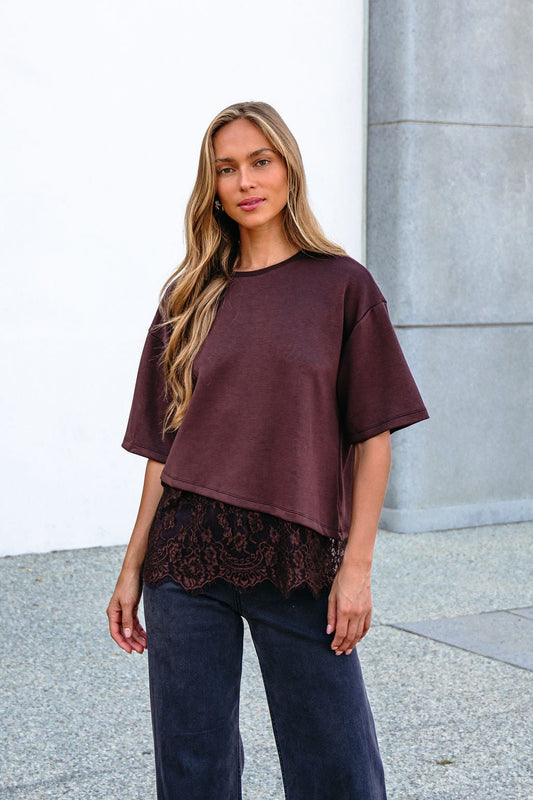 Woman in a Brown Lace Trim Short Sleeve Top over dark pants, standing outside by a light-colored wall.
