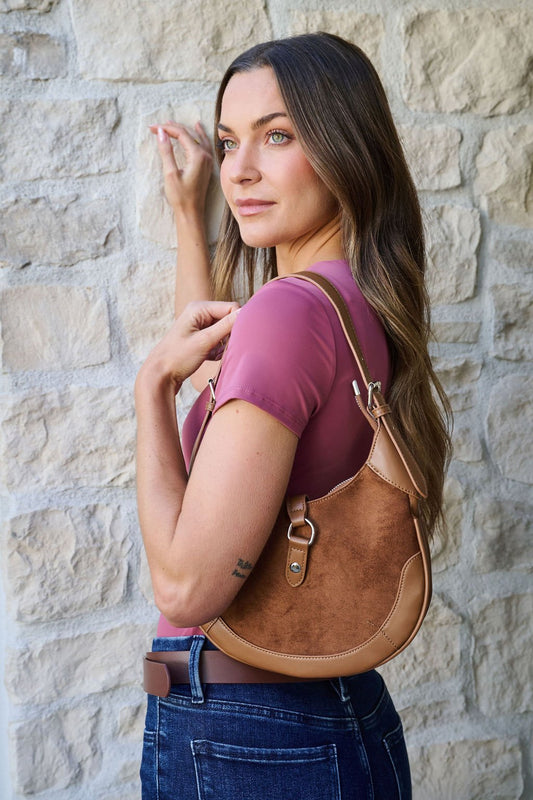 A woman in a mauve top and jeans stands by a stone wall, holding a Brown Leather Suede Crescent Shoulder Bag.