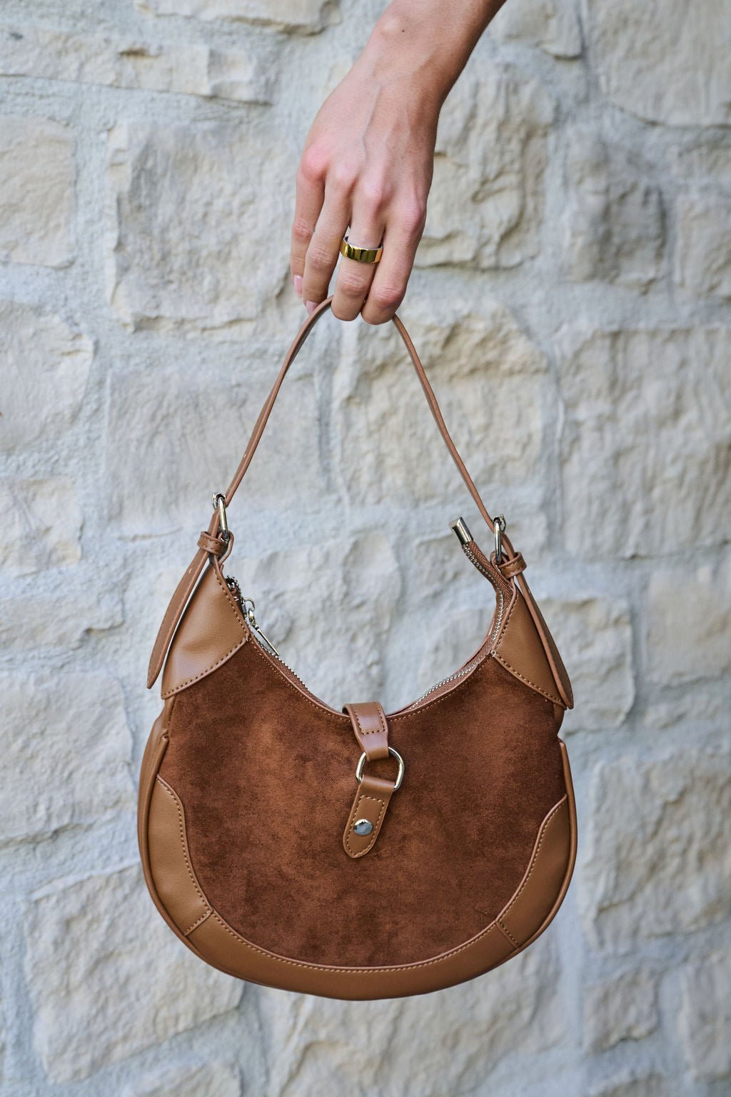 A hand holds the Brown Leather Suede Crescent Shoulder Bag against a light stone wall background.