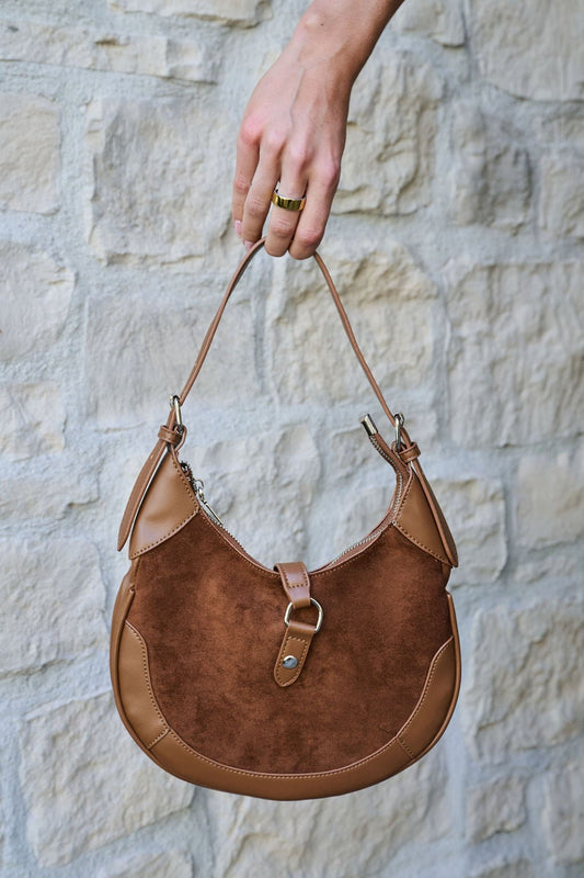 A hand holds the Brown Leather Suede Crescent Shoulder Bag against a light stone wall background.