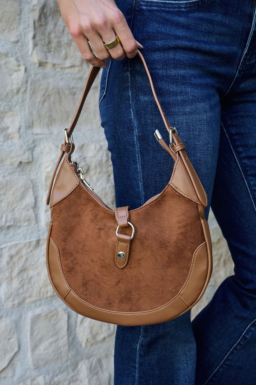 A person in jeans holds the Brown Leather Suede Crescent Shoulder Bag in front of a light stone wall.