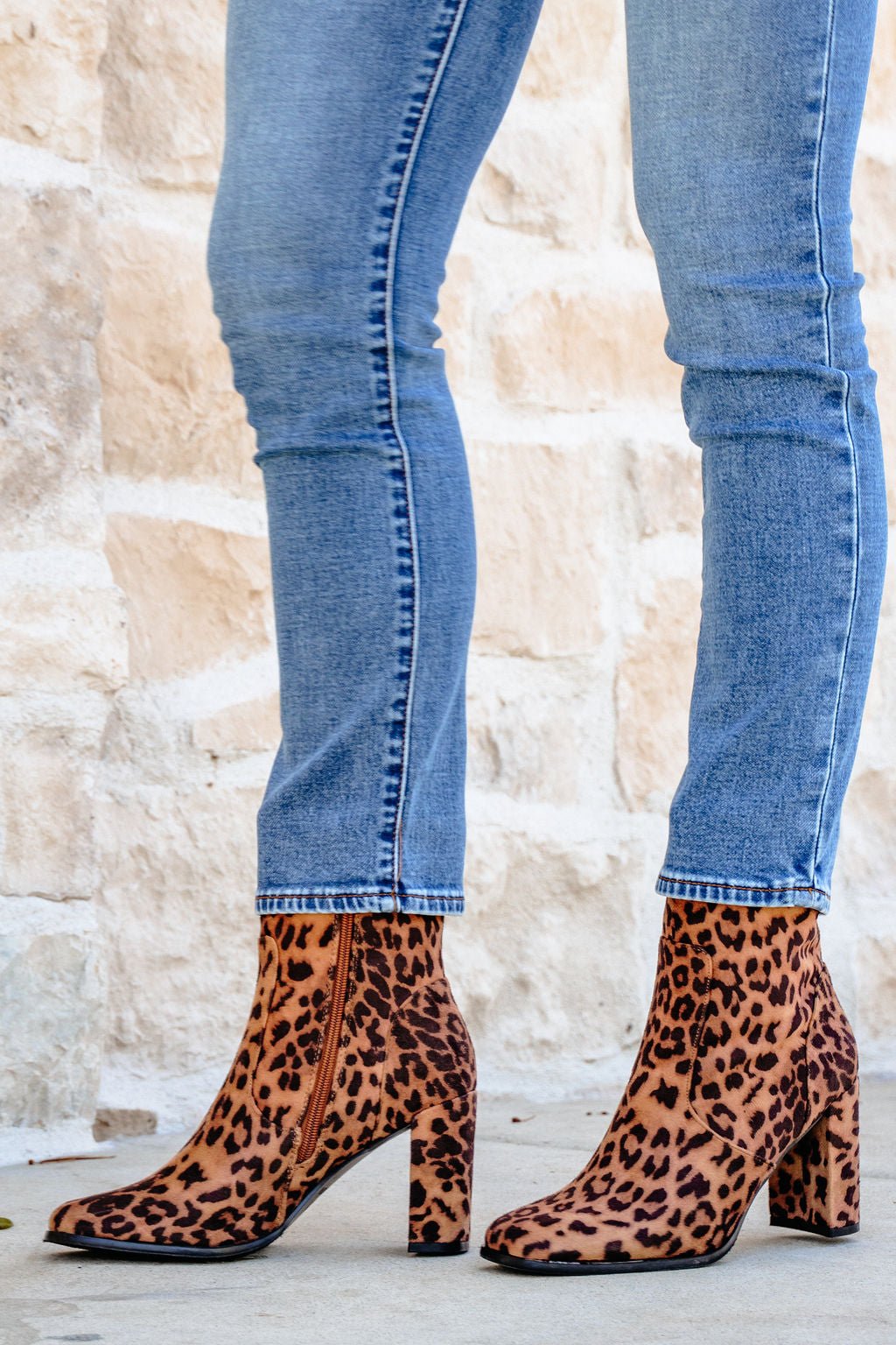 Person wearing blue skinny jeans and Brown Leopard Suede Booties, standing on light pavement near a stone wall.