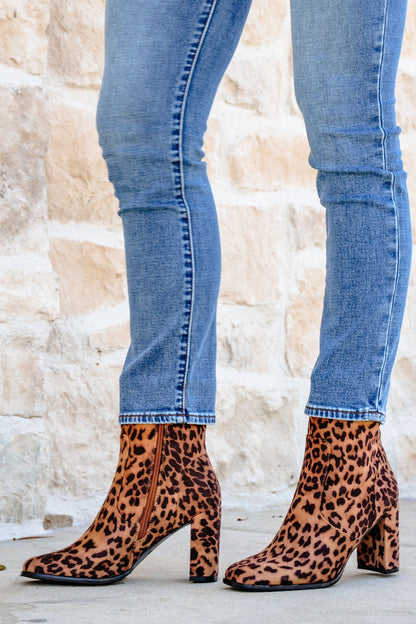 Person wearing blue skinny jeans and Brown Leopard Suede Booties, standing on light pavement near a stone wall.