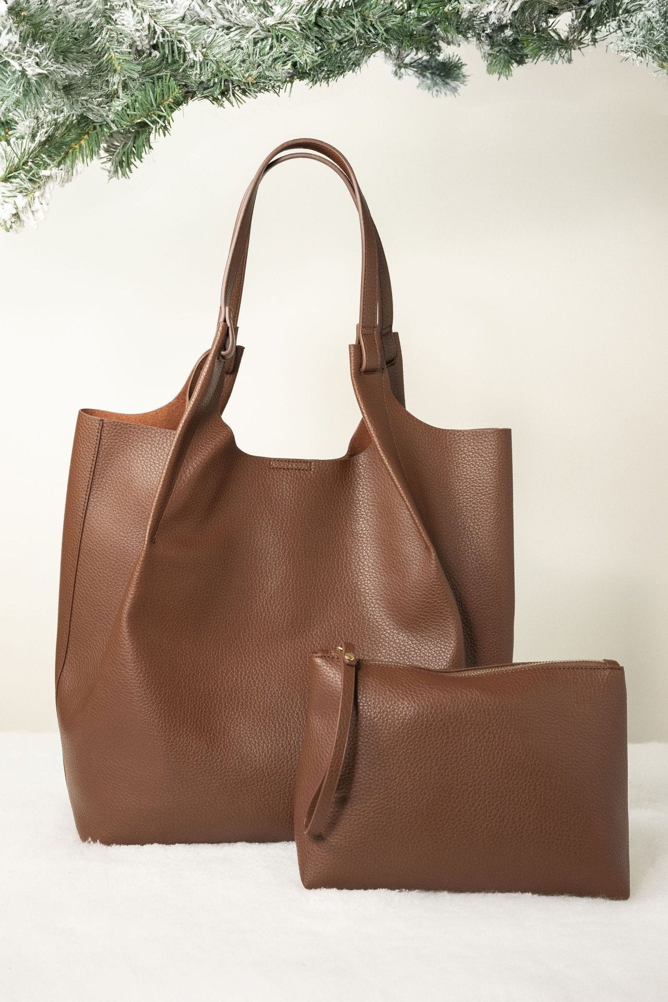 Brown Maeve Pebbled Leather Tote Bag with matching pouch, displayed on white surface with evergreen branches above.