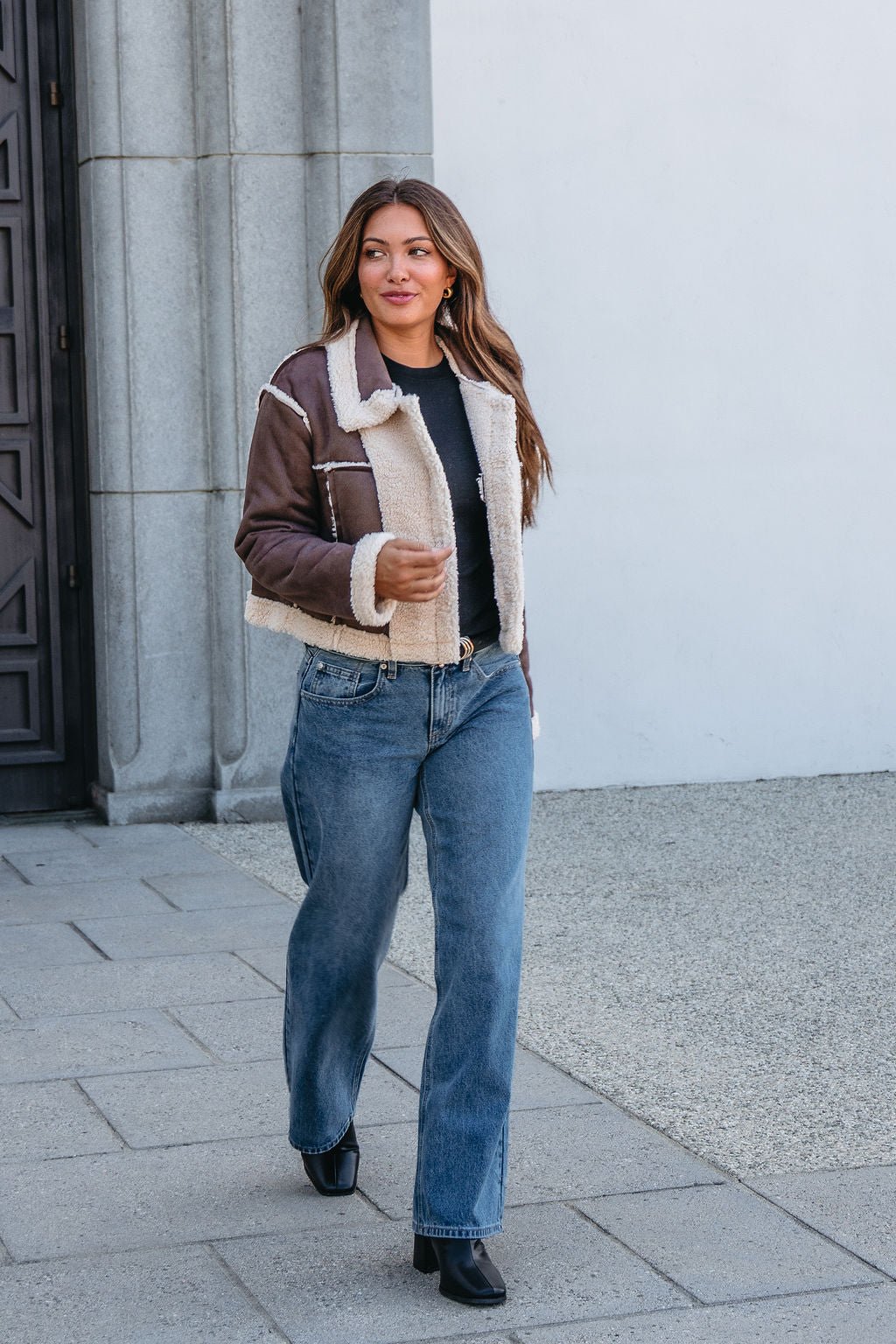 Woman wearing a Brown Mix Reversible Suede Sherpa Jacket, black top, blue jeans, and black boots walks on the sidewalk.