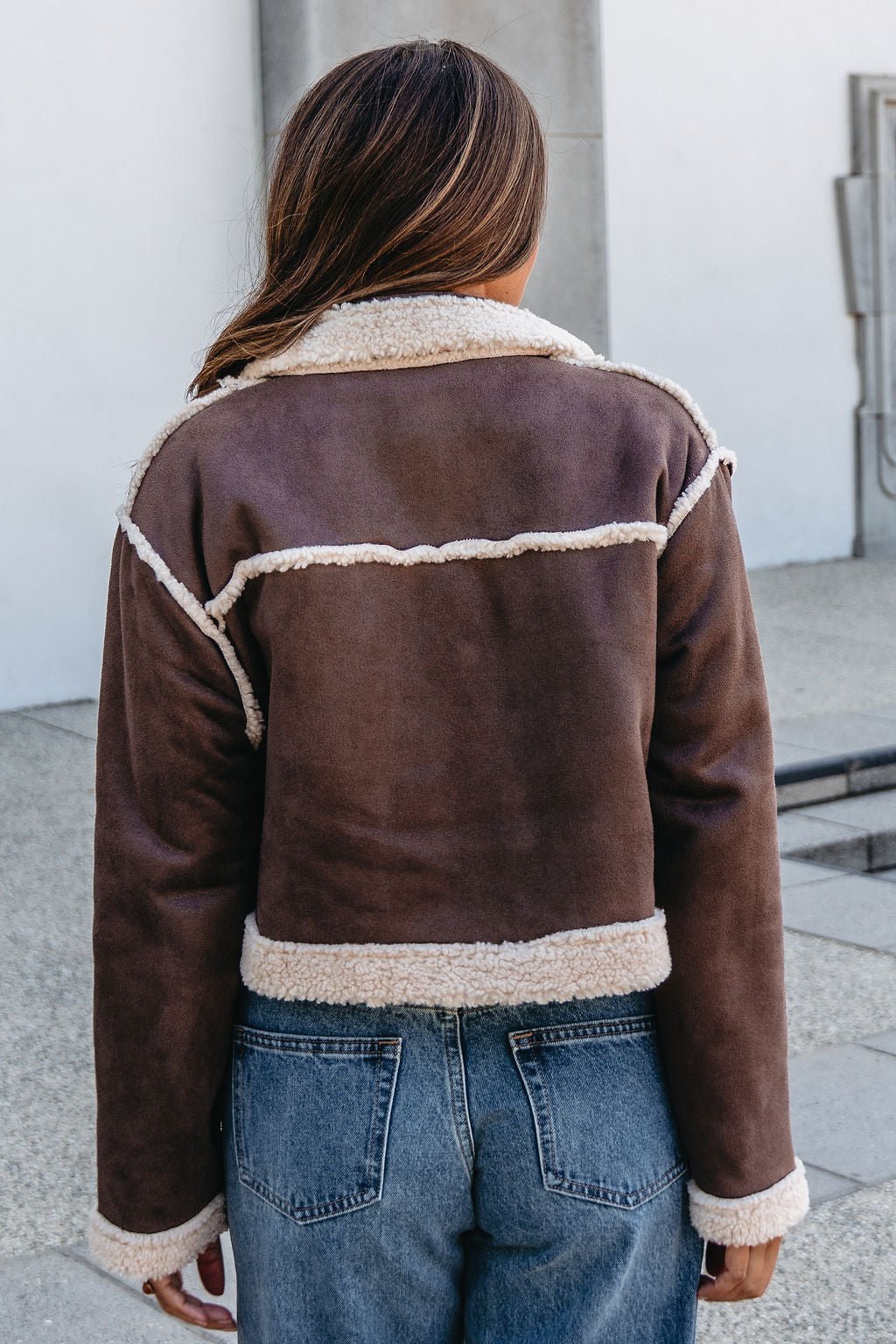 A woman outdoors, back to camera, wears a Brown Mix Reversible Suede Sherpa Jacket and blue jeans.