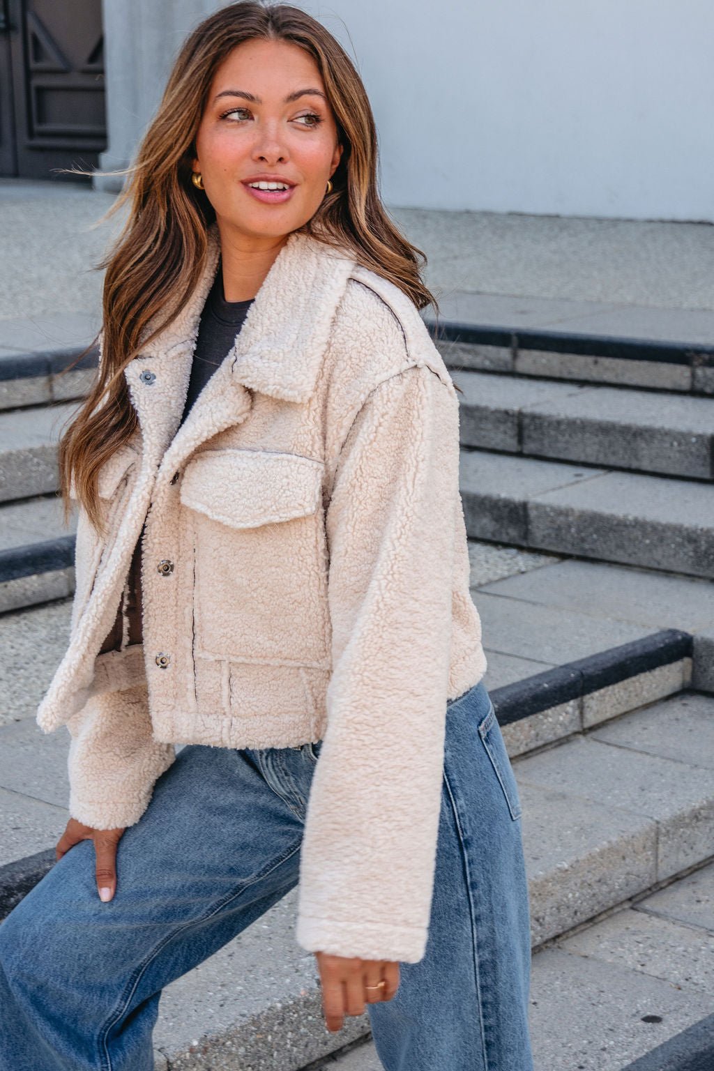 A woman with long hair wears a Brown Mix Reversible Suede Sherpa Jacket and blue jeans, sitting outdoors and looking to the side.