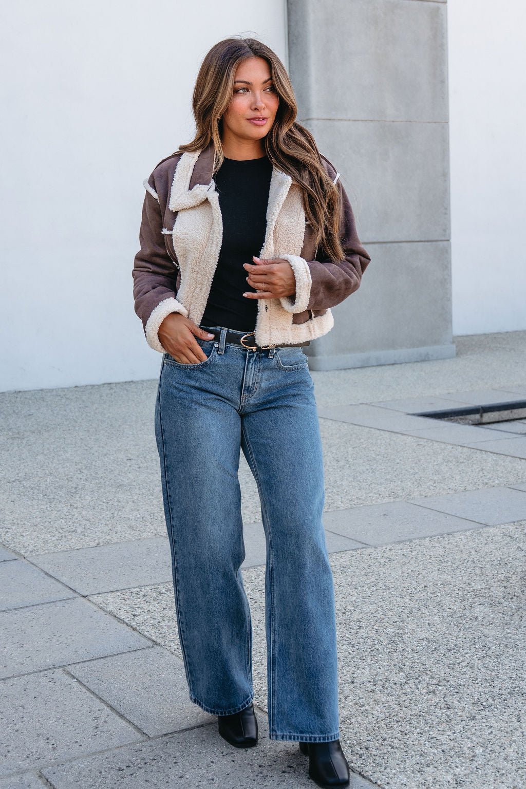 Woman in a Brown Mix Reversible Suede Sherpa Jacket, black top, jeans, and boots walks outdoors on a paved surface.