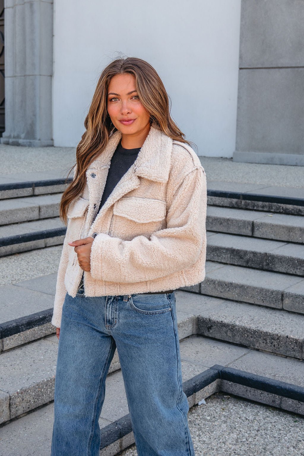 A woman wears a Brown Mix Reversible Suede Sherpa Jacket and blue jeans, standing outdoors on stone steps, smiling at the camera.