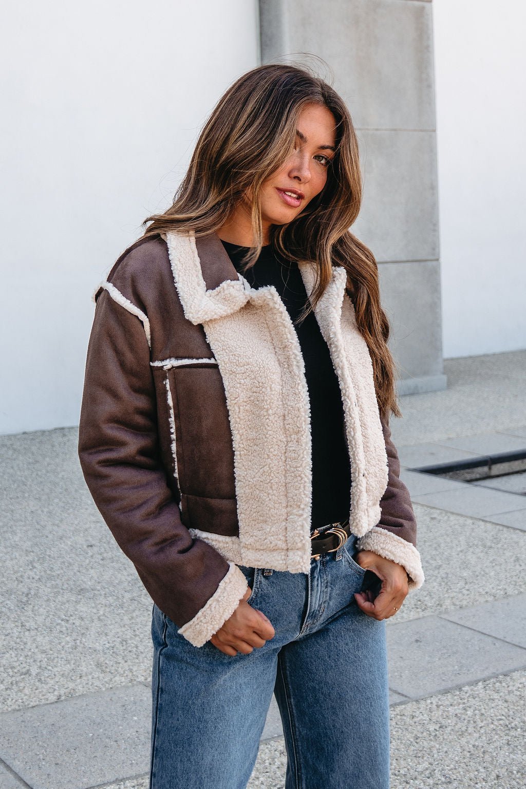 A woman in a Brown Mix Reversible Suede Sherpa Jacket, black top, and blue jeans stands outside by a light wall.
