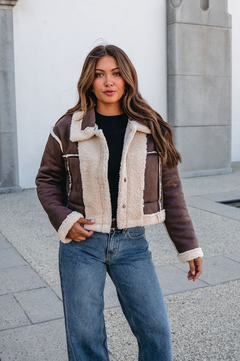 A woman in a Brown Mix Reversible Suede Sherpa Jacket, black top, and jeans stands outside on a stone walkway, facing the camera.