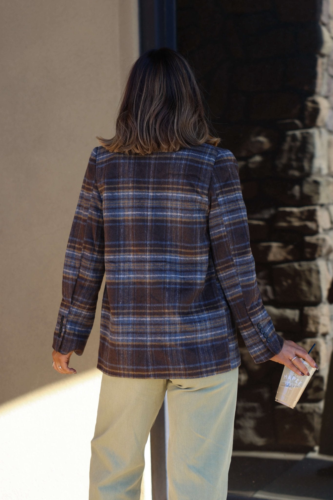 Person with shoulder-length brown hair in a Brown Plaid Blazer Jacket and light pants, holding a drink, facing away from camera.