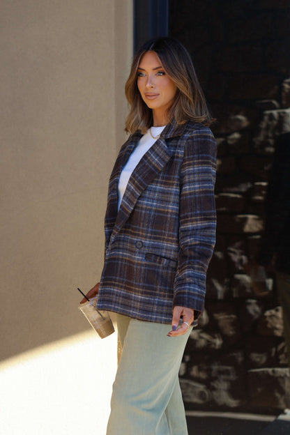 A woman in a Brown Plaid Blazer Jacket and light pants holds a drink cup outdoors, wearing a neutral expression.