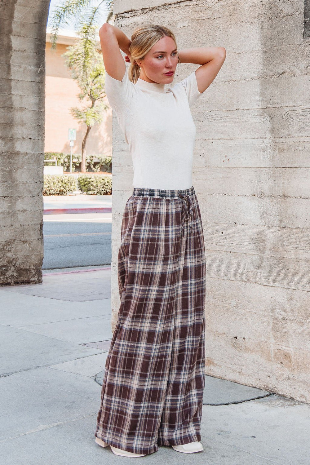 Woman on the sidewalk wearing Brown Plaid Wide Leg Pants - FINAL SALE, tying her hair back.