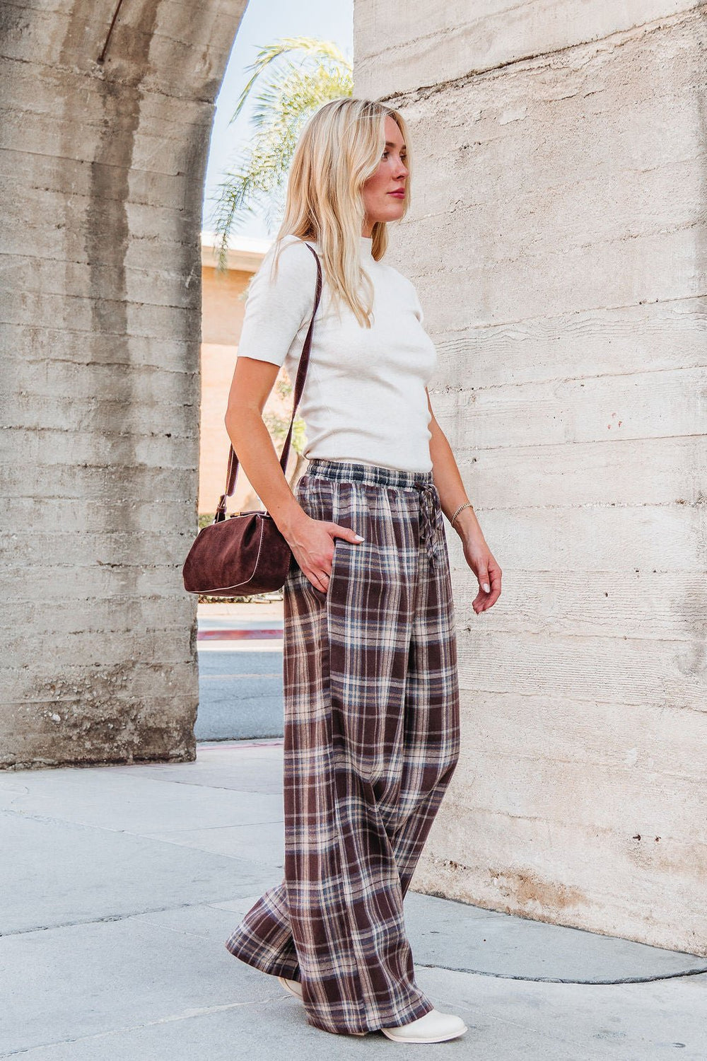 Woman in Brown Plaid Wide Leg Pants - FINAL SALE, white top and shoes, brown bag, walks by a concrete wall.