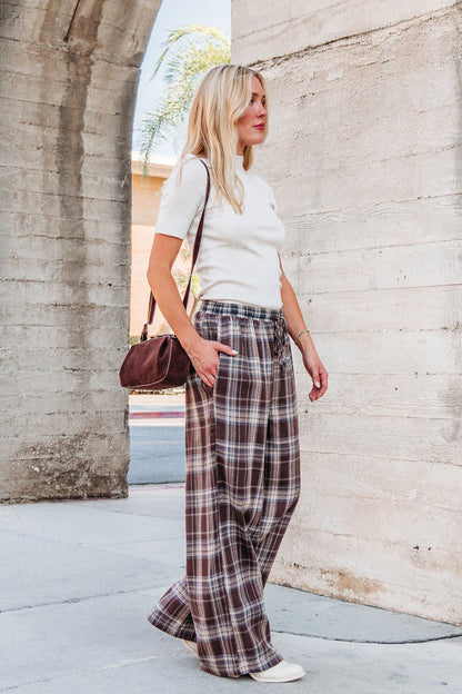 A woman stands outside wearing Brown Plaid Wide Leg Pants - DOORBUSTER, a white top, white shoes, and a brown shoulder bag.