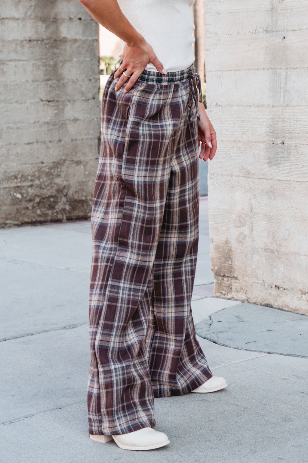 Person wearing Brown Plaid Wide Leg Pants - DOORBUSTER, white shoes, and a white top stands by a concrete wall outdoors.