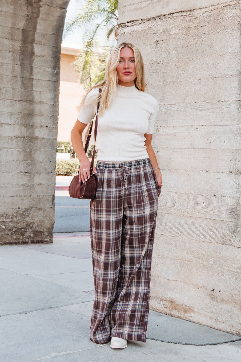 Woman stands outside in Brown Plaid Wide Leg Pants - DOORBUSTER, white short-sleeve top, white shoes, and a small brown bag.