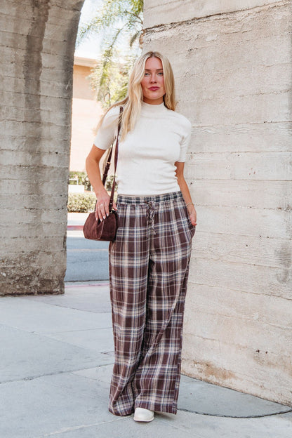 Woman stands outside in Brown Plaid Wide Leg Pants - DOORBUSTER, white short-sleeve top, white shoes, and a small brown bag.