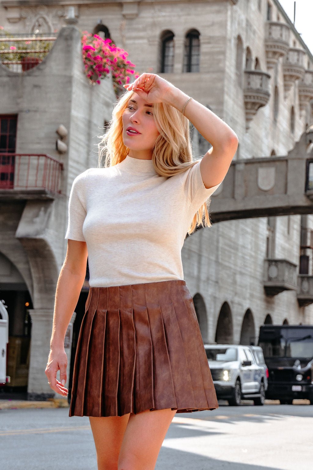 A woman wearing a Brown Pleated Faux Leather Mini Skirt stands on a sunny city street, shielding her eyes with her hand.