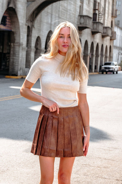 A woman in a beige top and Brown Pleated Faux Leather Mini Skirt stands on a sunlit city street near stone arches.