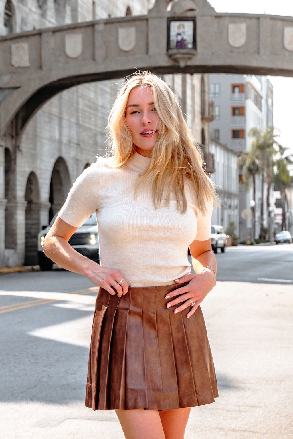 A woman in a white top and Brown Pleated Faux Leather Mini Skirt stands confidently on a sunny city street with historic buildings.