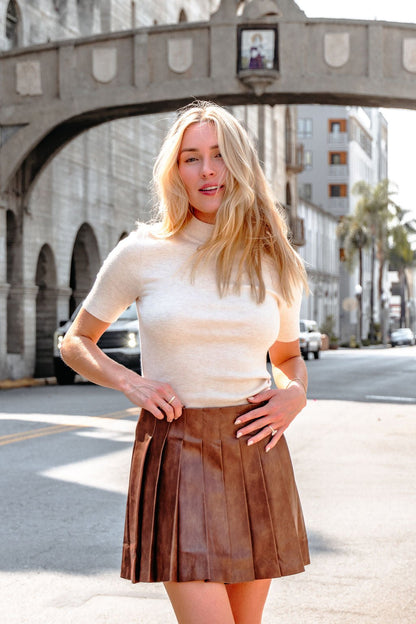 A woman in a white top and Brown Pleated Faux Leather Mini Skirt stands confidently on a sunny city street with historic buildings.