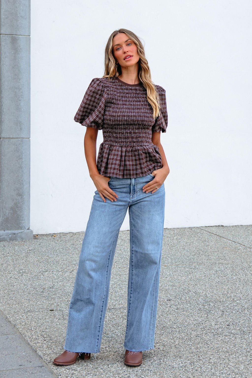 Woman in a Brown Puff Sleeve Peplum Top and wide-leg jeans stands outdoors, posing with hands in her pockets.