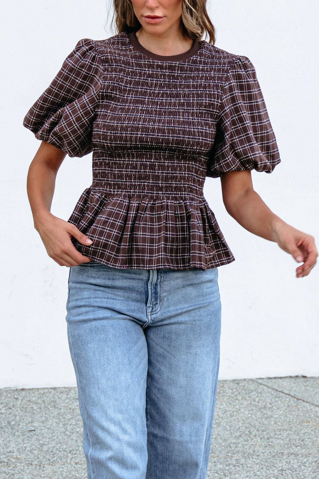 Woman wears a Brown Puff Sleeve Peplum Top with light blue jeans, standing outdoors against a plain white background.