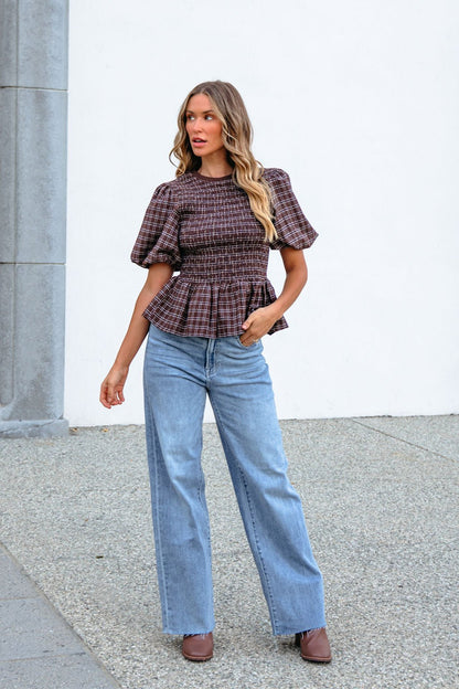 Woman in a brown puff sleeve peplum top, wide-leg jeans, and brown shoes stands on pavement by a gray column and white wall.