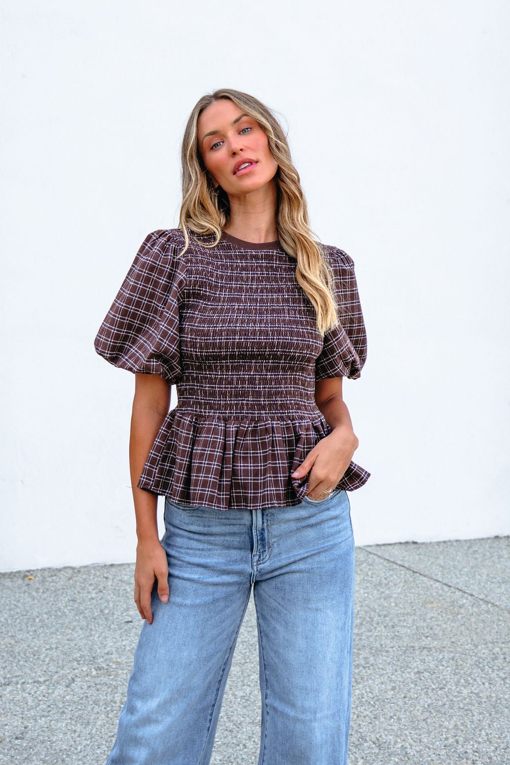 Woman wears a Brown Puff Sleeve Peplum Top with blue jeans, posing in front of a white wall and looking at the camera.
