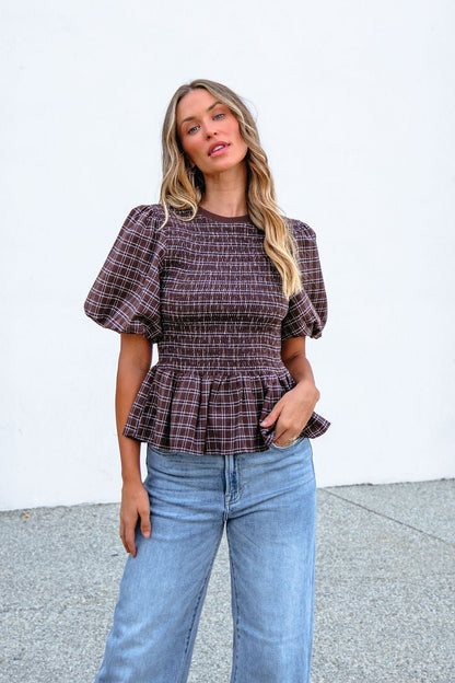 Woman wears a Brown Puff Sleeve Peplum Top with blue jeans, posing in front of a white wall and looking at the camera.