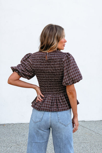 A woman faces away in a Brown Puff Sleeve Peplum Top and light blue jeans, set against a plain white wall.