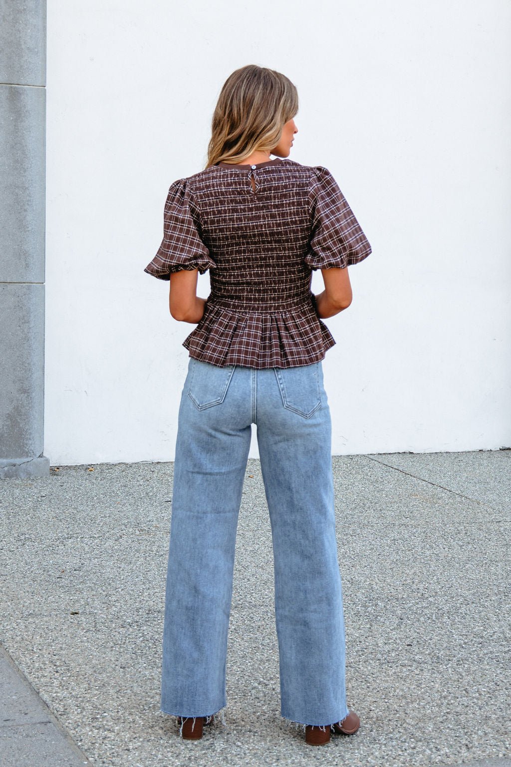 A woman stands outdoors facing away, in a brown puff sleeve peplum top and light blue wide-leg jeans.