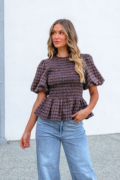 Woman models the Brown Puff Sleeve Peplum Top with light blue jeans against a white wall, glancing to the side.