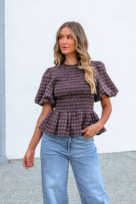 Woman models the Brown Puff Sleeve Peplum Top with light blue jeans against a white wall, glancing to the side.