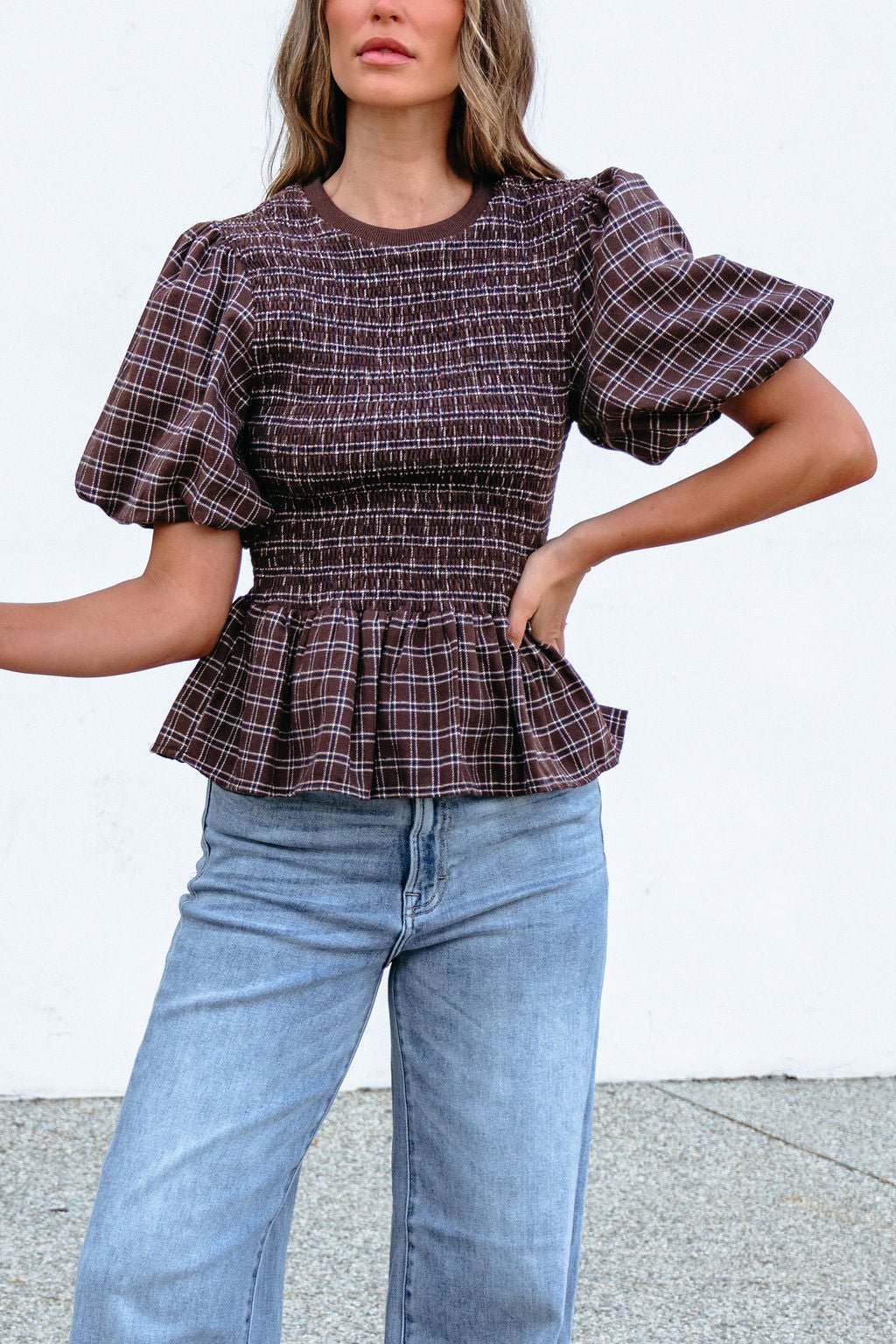 Woman wears a Brown Puff Sleeve Peplum Top with light blue jeans, standing against a plain background.