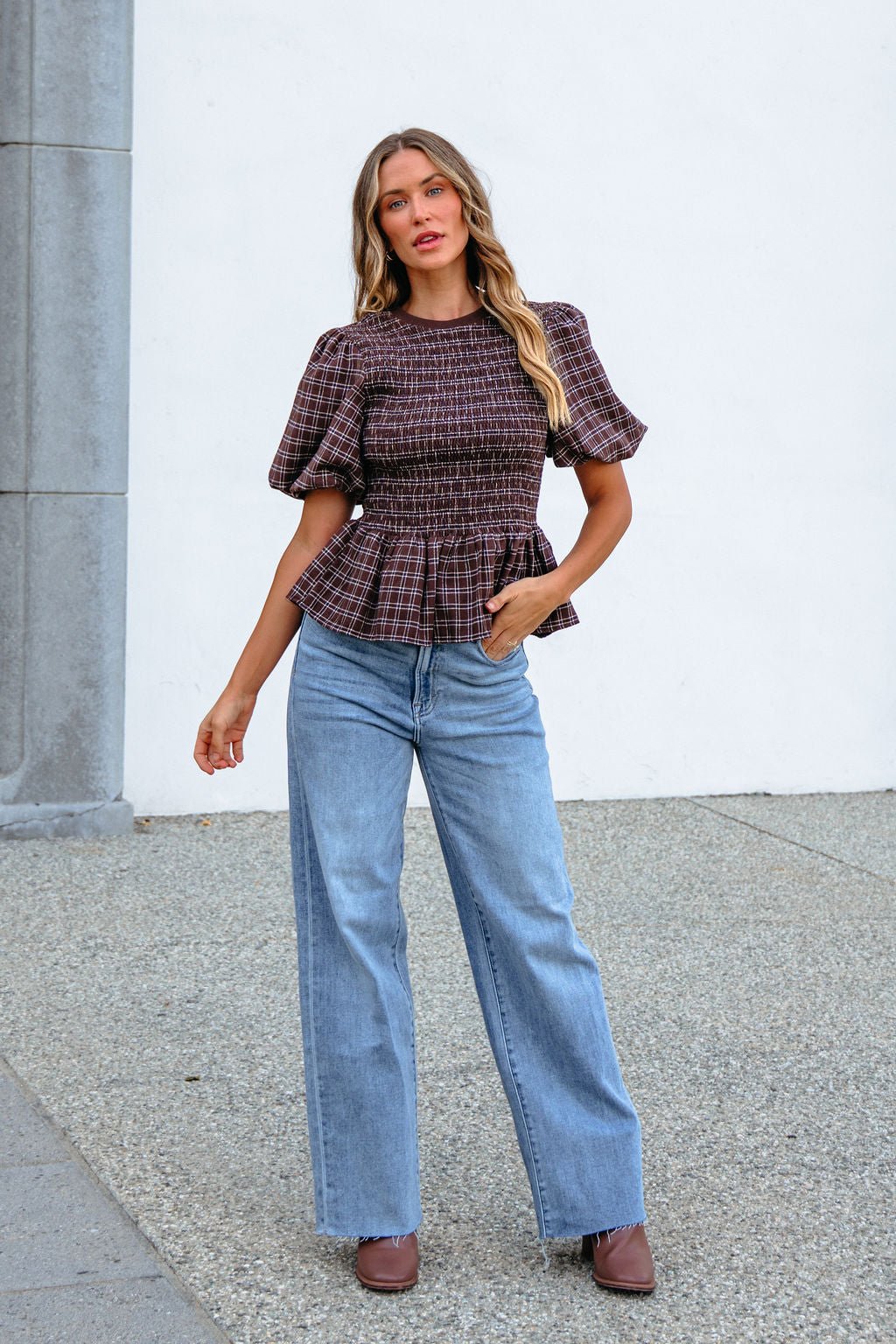 Woman wearing a Brown Puff Sleeve Peplum Top and wide-leg jeans poses outside, one hand on her hip.