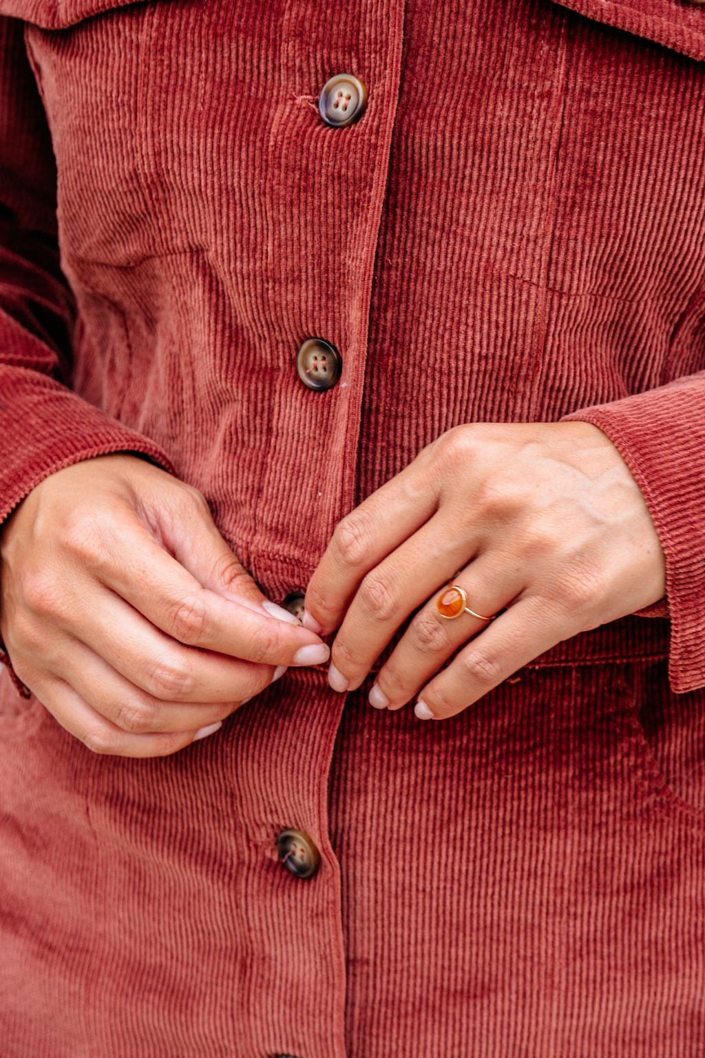 A person in a red corduroy jacket buttons up, showcasing the Brown Resin Acrylic Band Statement Ring - DOORBUSTER.
