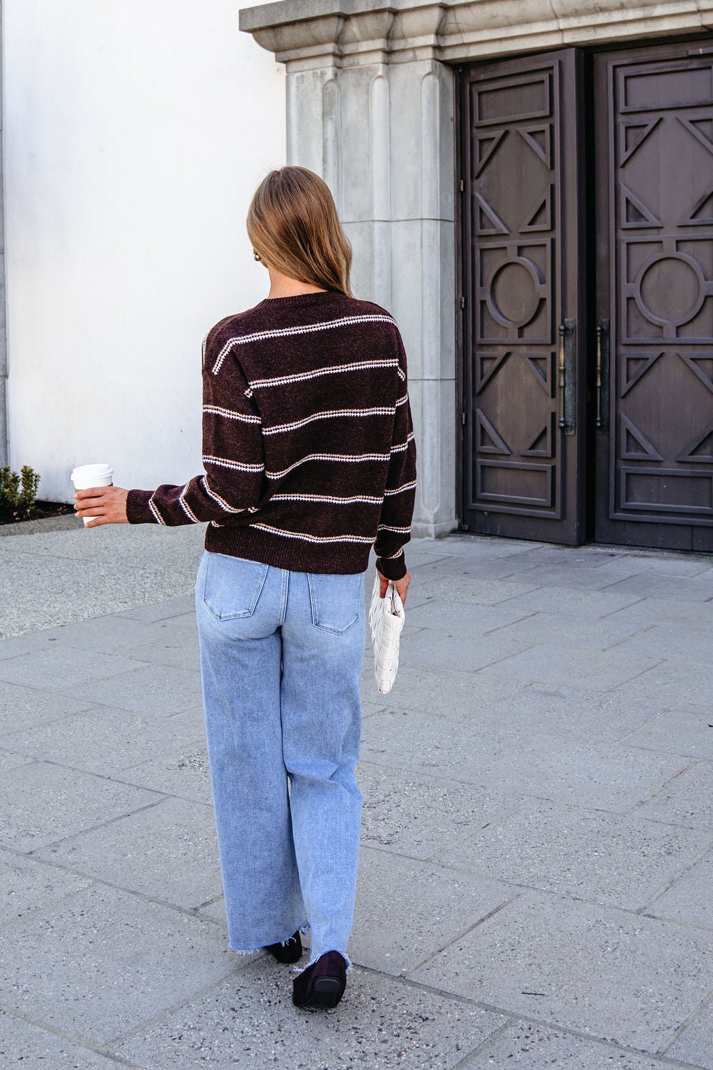 Someone wearing a Brown Rib Knit Striped Sweater and light blue jeans walks outside with a coffee cup, facing away from the camera.