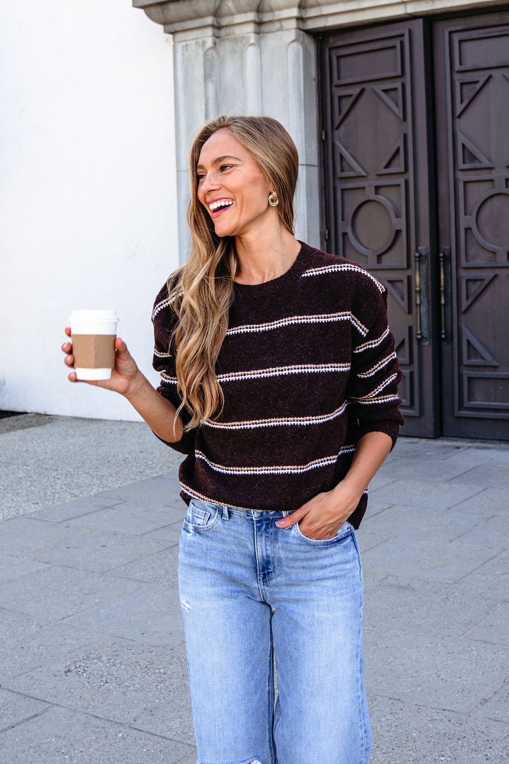 Woman smiles outdoors holding coffee, wearing the Brown Rib Knit Striped Sweater with jeans in front of a large door.