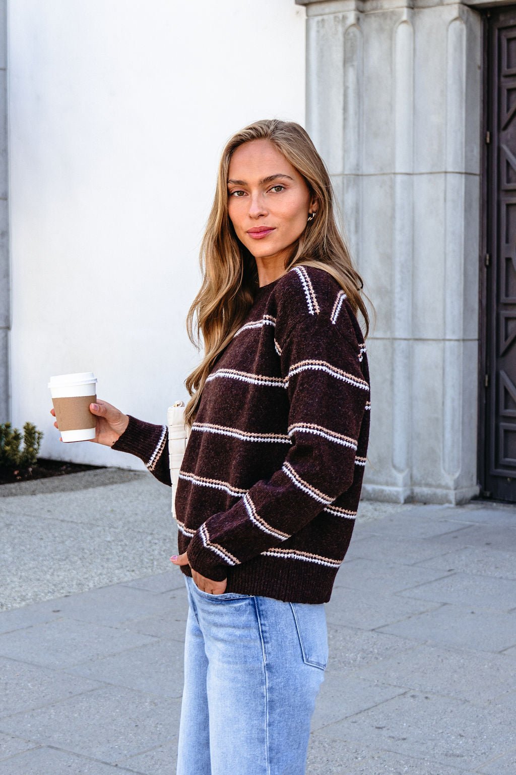 Woman in a Brown Rib Knit Striped Sweater and jeans holds a coffee cup outdoors by a stone building, looking at the camera.