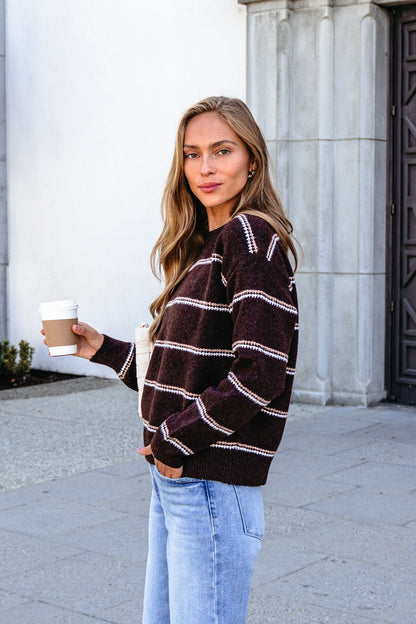 Woman in a Brown Rib Knit Striped Sweater and jeans holds a coffee cup outdoors by a stone building, looking at the camera.