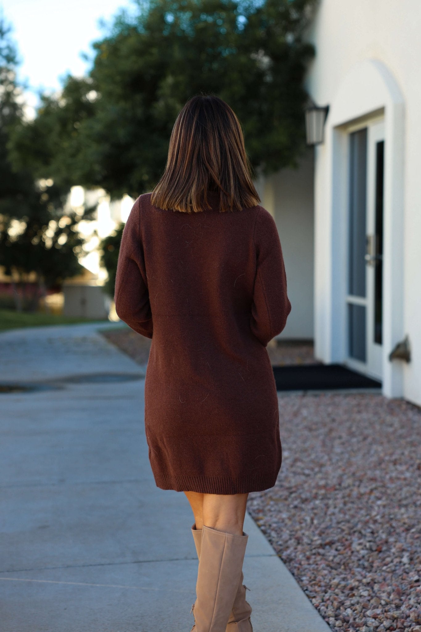 A woman in a Brown Ribbed Cardigan Sweater Dress and tan boots walks on a sidewalk by a white building.