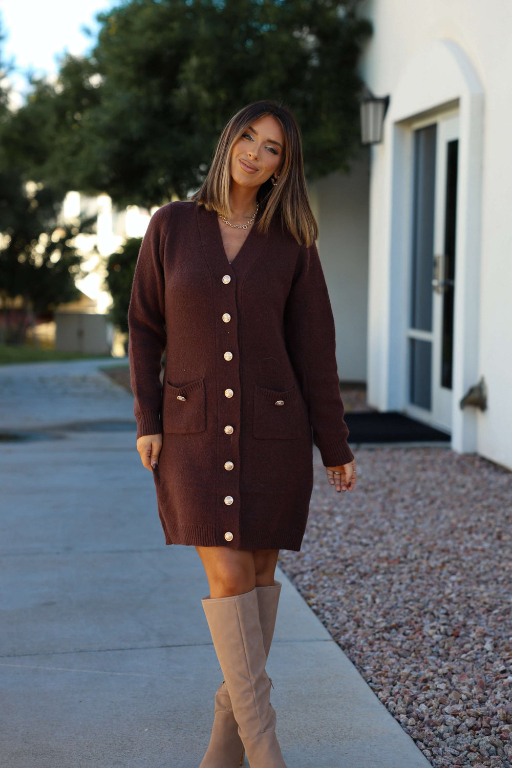 A woman in the Brown Ribbed Cardigan Sweater Dress smiles outside near a white building, wearing beige knee-high boots.