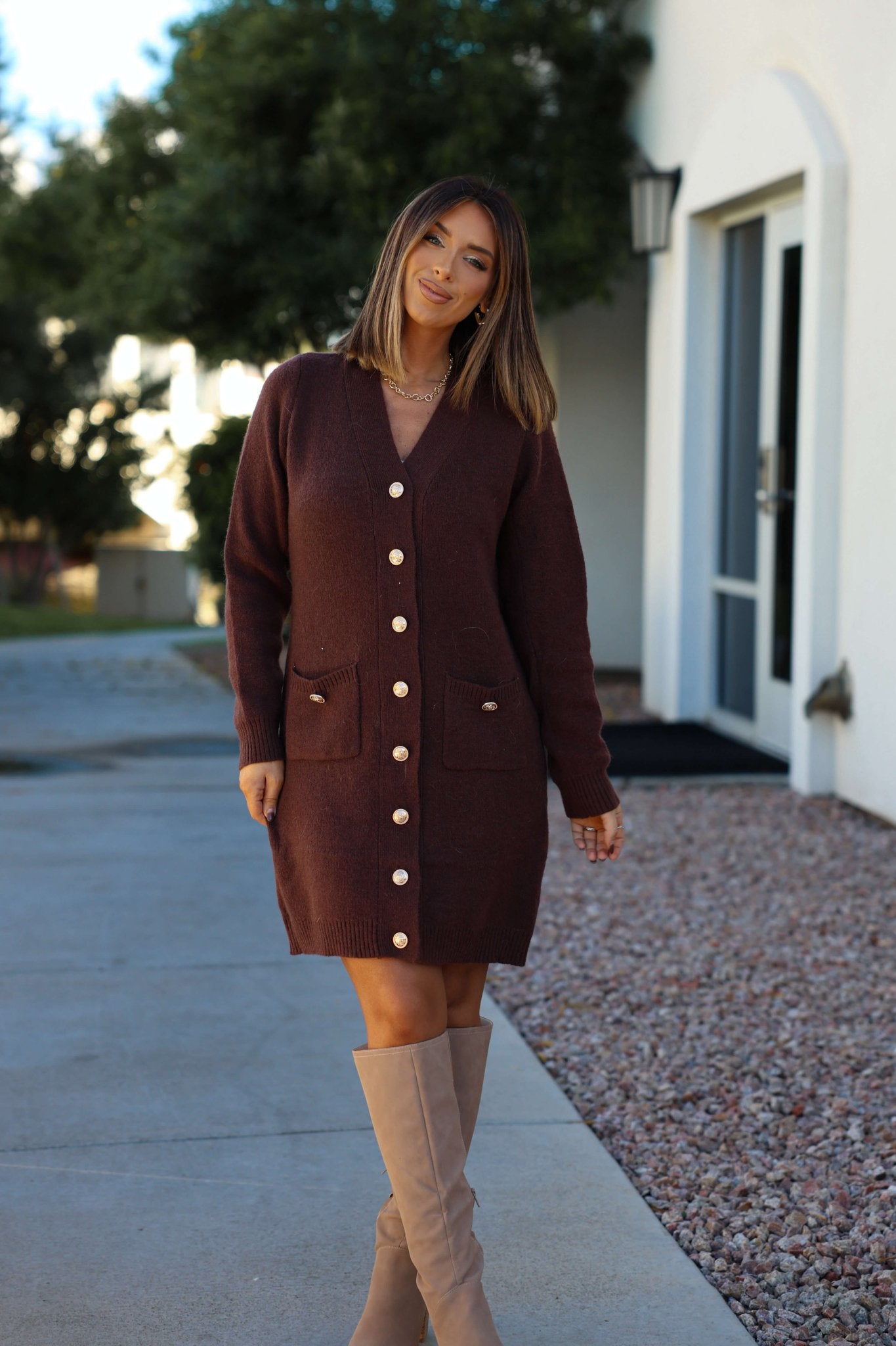 A woman in a Brown Ribbed Cardigan Sweater Dress smiles outdoors by a white building, wearing beige knee-high boots.