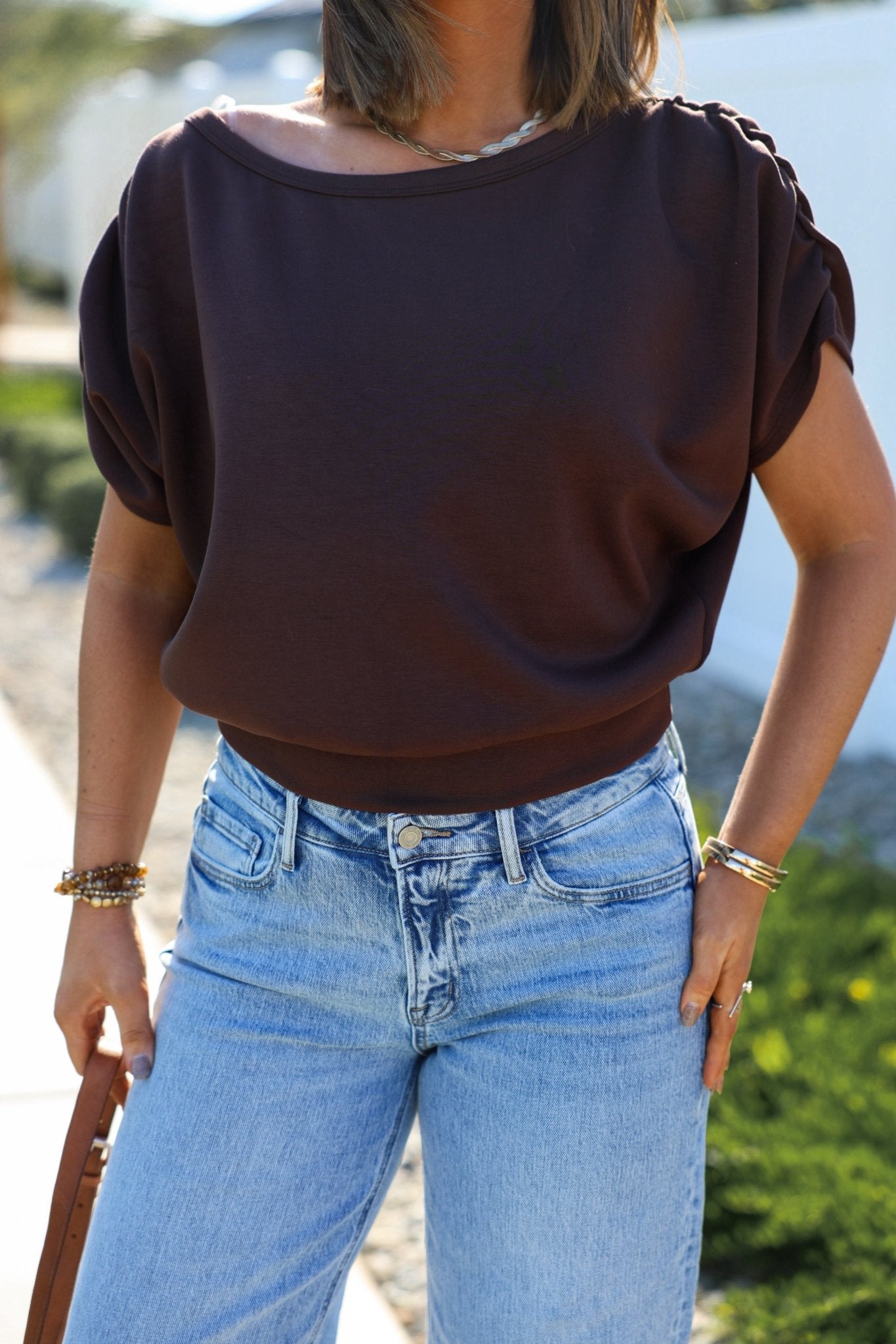 Outdoors, a woman in a Brown Shirred Sleeve Boatneck Top and jeans holds a brown bag; her look is effortlessly American-made.