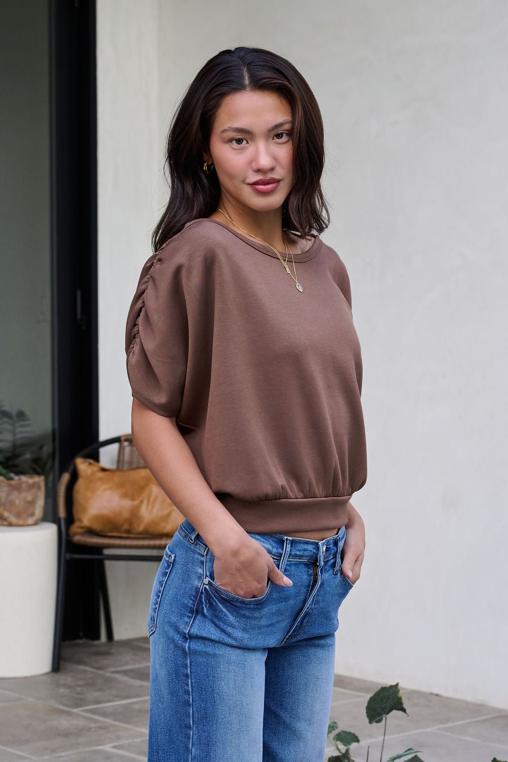 A woman wearing a Brown Shirred Sleeve Boatneck Top and blue jeans stands outdoors, hands in pockets, facing the camera.