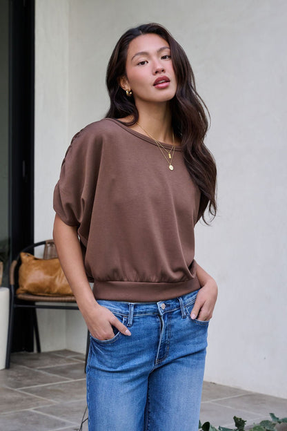 A woman wears a Brown Shirred Sleeve Boatneck Top and blue jeans, standing outdoors with confidence and one hand in her pocket.